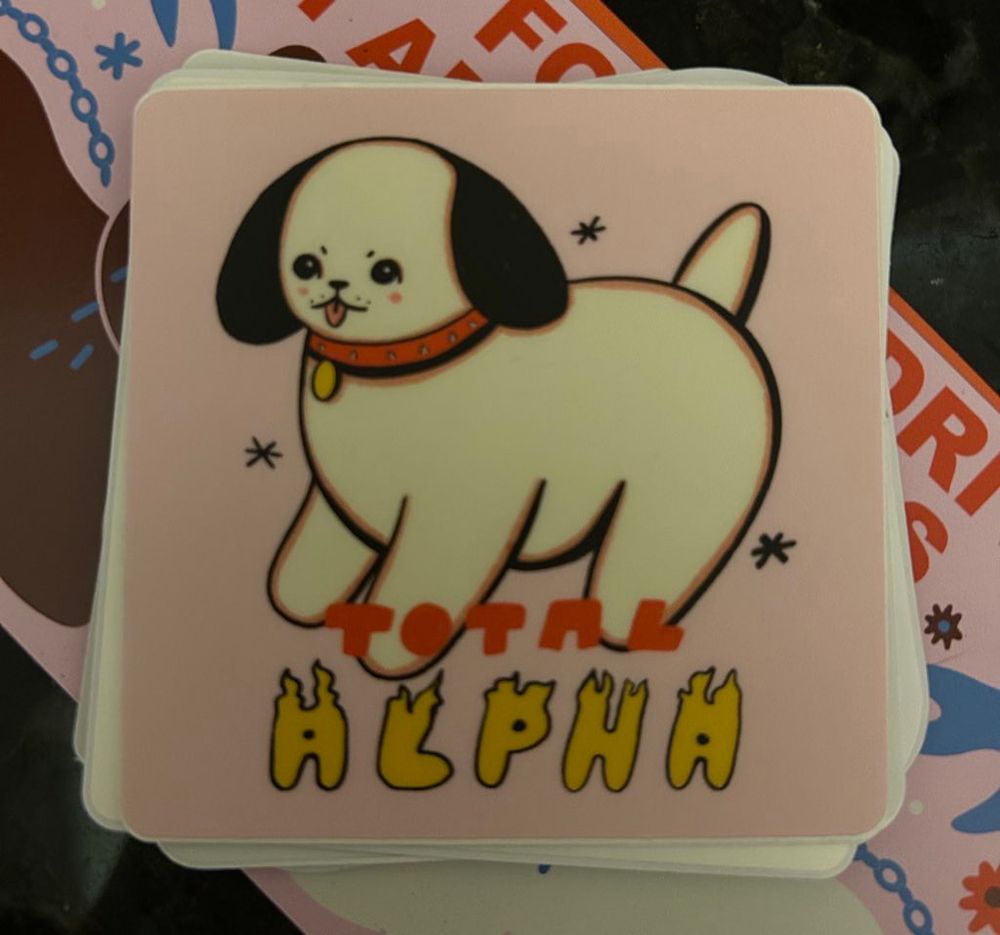 Photo of a sticker by Tender Ghost of a round little dog, white with black ears, with a red collar and his tongue out that says TOTAL ALPHA on the bottom