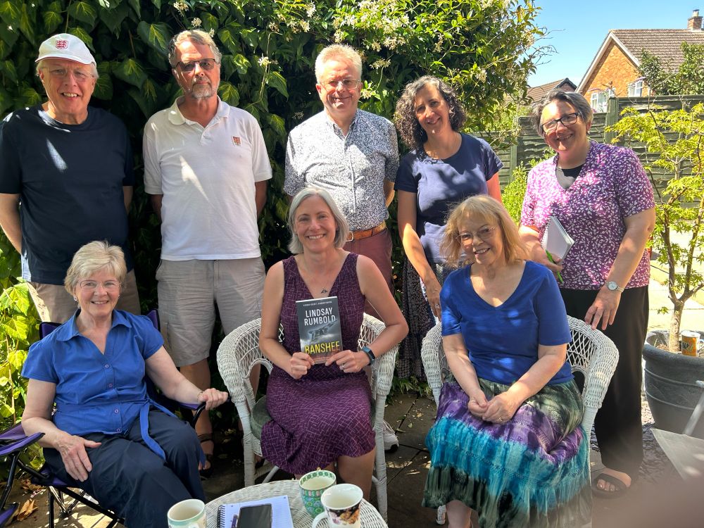 Me, a 40 something woman in a purple dress with jaw length grey hair, sitting between Heather Flack (L) and Sheila aka SC Skillman (R). Behind us stand 5 more people, including Nigel Oakley (centre) and Fran Hill (R)