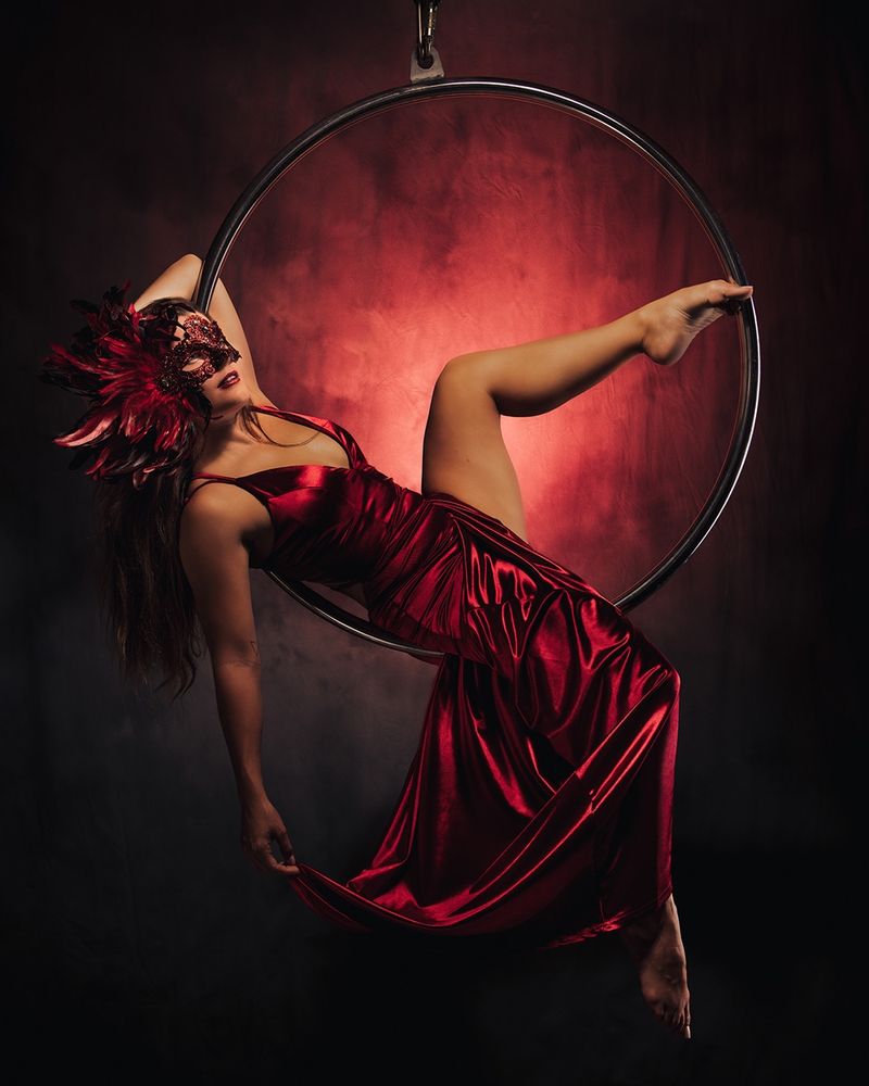 Brunette model in a red and black jeweled and feathered mask, and a long red gown.  She is laying in a lyra hoop, dangling in mid-air.