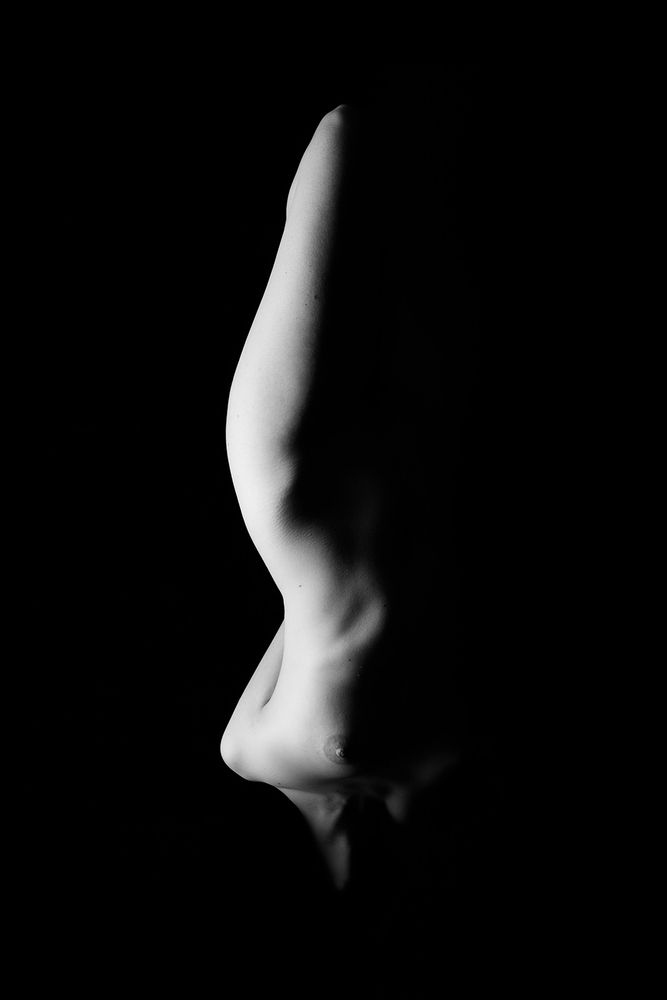 Black and white, low key image of a nude woman, one side of her body visible from her knee to her neck with the other side falling off into darkness.  Her form is inverted from our viewpoint, and her head and lower legs are obscured in darkness, effects which combine to create both a familiar and beautiful, while also unsettling and abstract image.