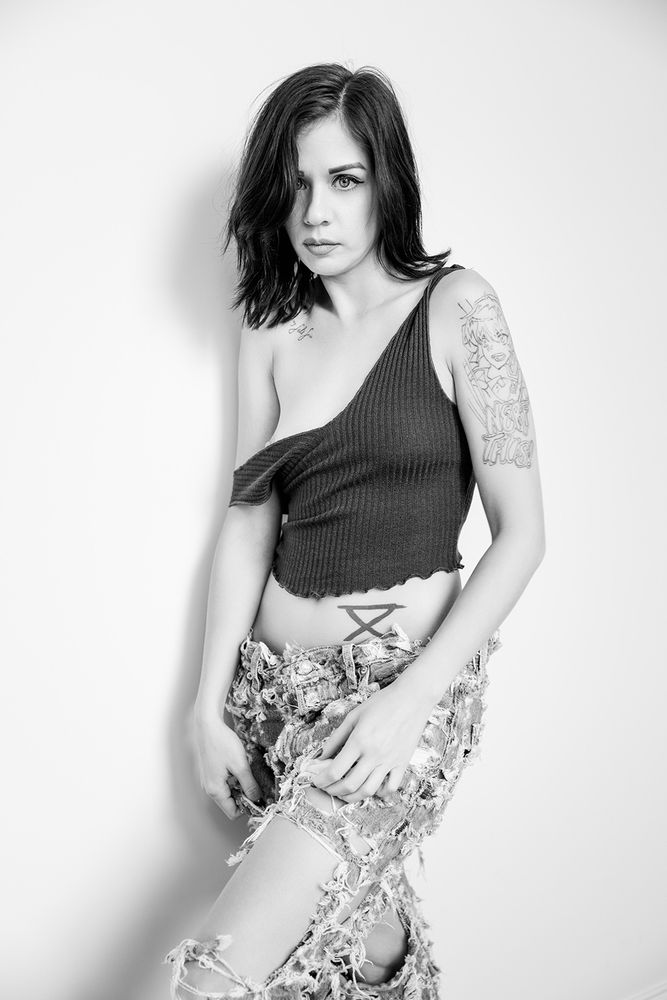 B&W image of a brunette model in torn up denim jeans and a dark colored tank top.  The tank top has slipped off of one shoulder, partially exposing her nipple.