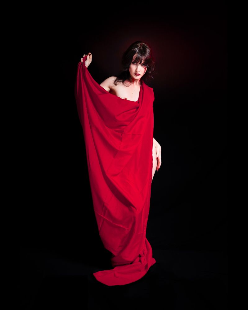 Low key image of a brunette model wrapped in a red sheet, which has dropped just a bit in the front to expose one breast.  She stands against a black background which is ever so slightly lit with red.