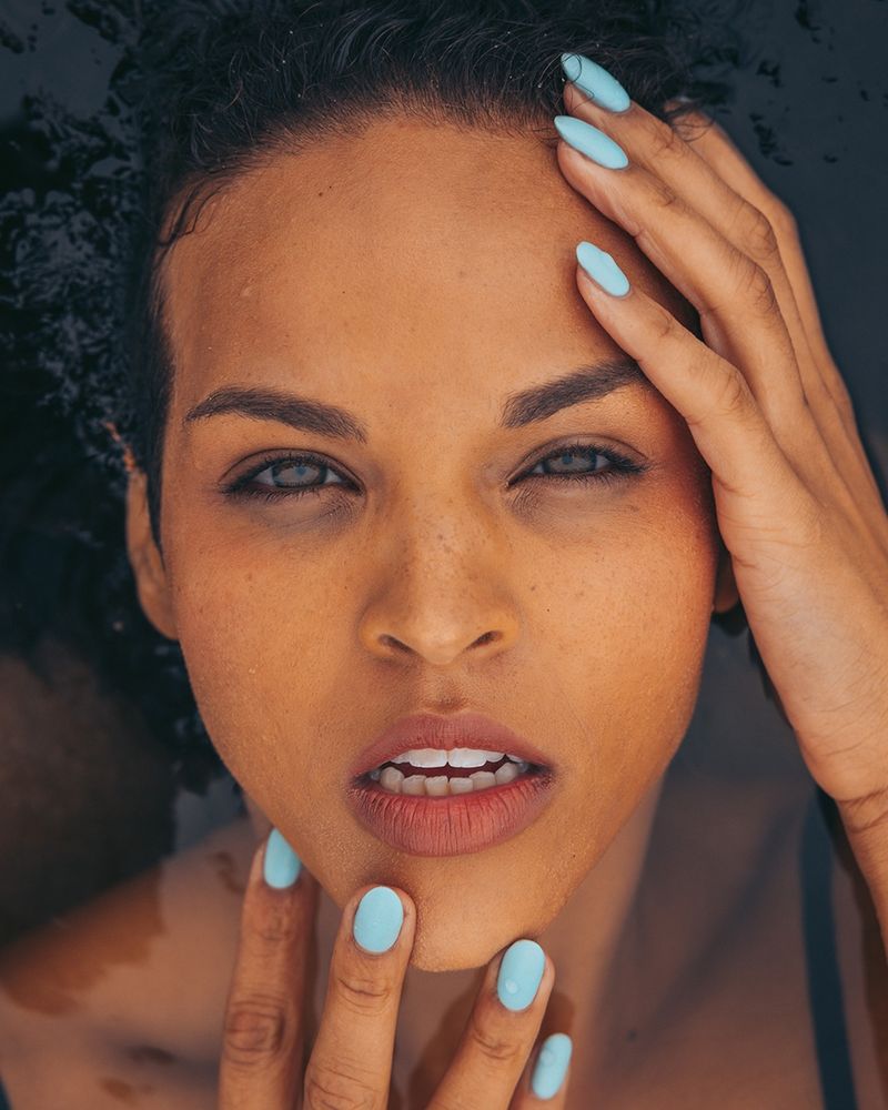Portrait of a brown-eyed, brunette model laying in a tidal pool, her hair melting away into the water which is also covering her body.  Her hands, tipped with light blue fingernails, frame her face.