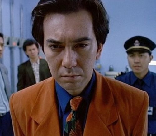 Anthony Wong in a  bright orange suit with a blue shirt and a rose print tie.