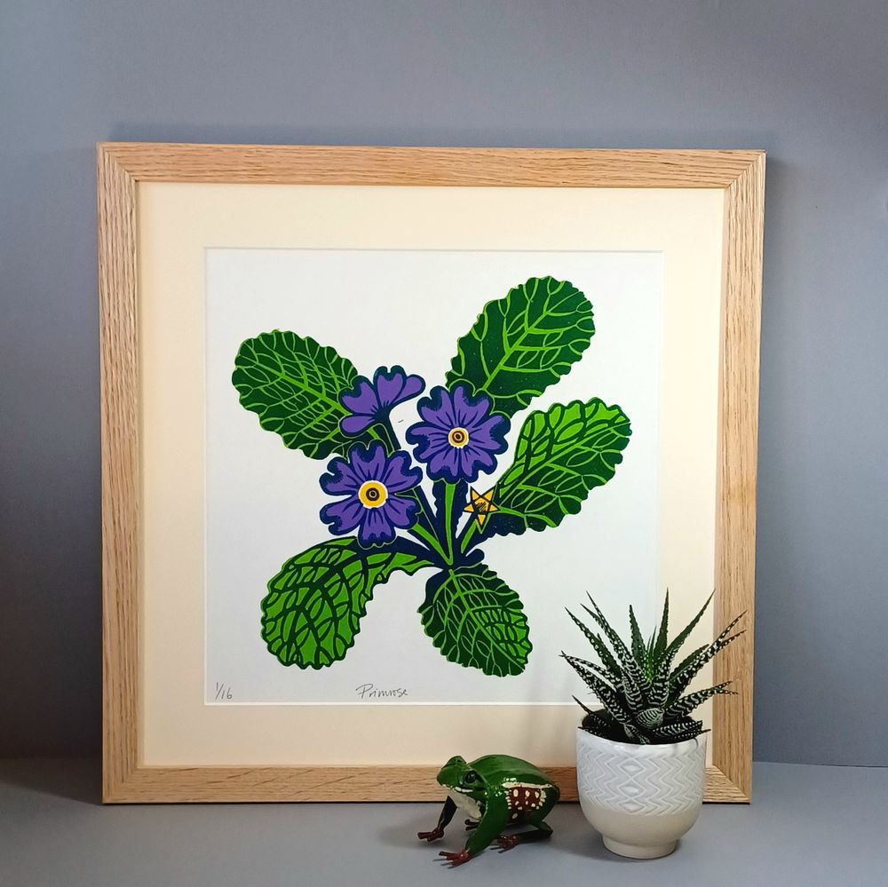 Framed linocut print of a flowering purple primrose with patterned leaves. In front of the print is a potted plant and frog ornament. The background is grey.