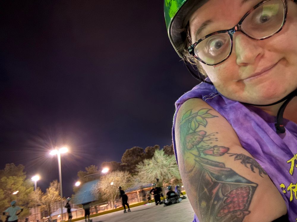 A woman in a watermelon print helmet with a bat tattoo on her upper arm smiles, while people roller skate behind her 