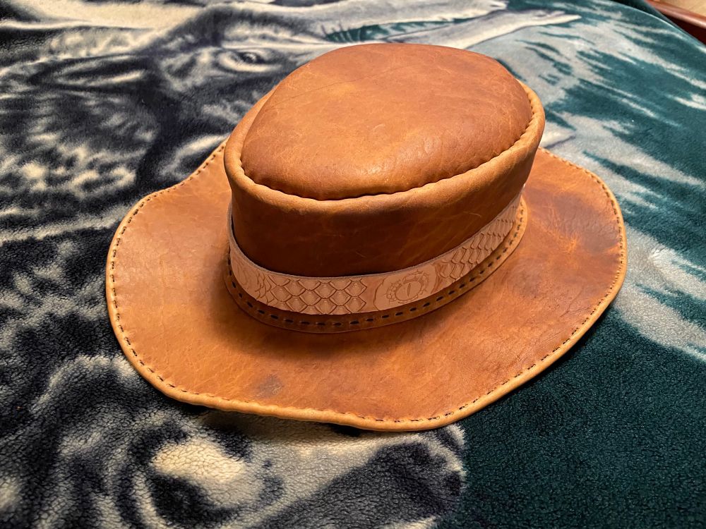 Front three-quarter view of a wide-brimmed sand-colored hat. The hat band has thick scales and a draconian eye stamped into it. 