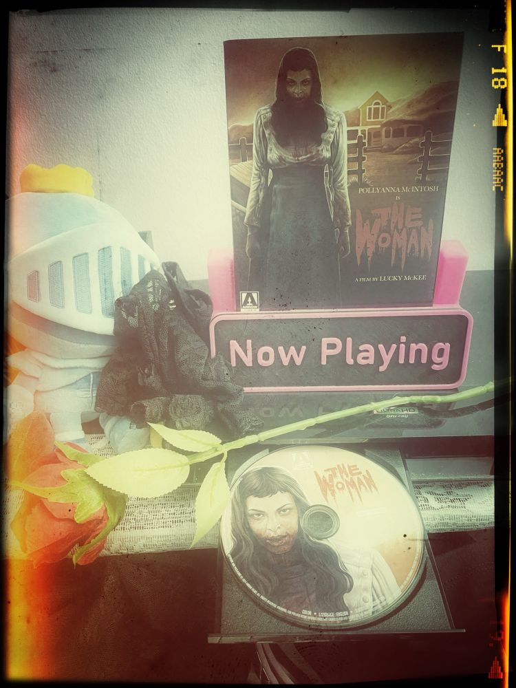 Case for “The Woman“ Arrow edition Blu-ray in a pink and black ‘now playing’ stand on top of a Blu-ray player. Disc is in the tray.

There’s also a plastic rose in front of the now playing stand to add drama.
