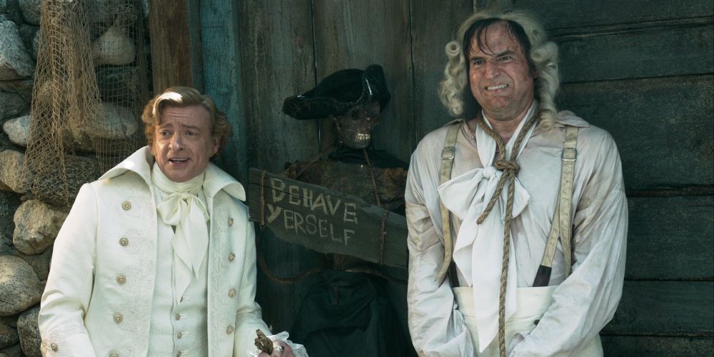 Stede and Hornsberry standing on either side of a skeleton wearing a tricorn pirate hat and holding a sign that says “Behave Yerself”. Hornsberry is doing his best to “look tortured”.