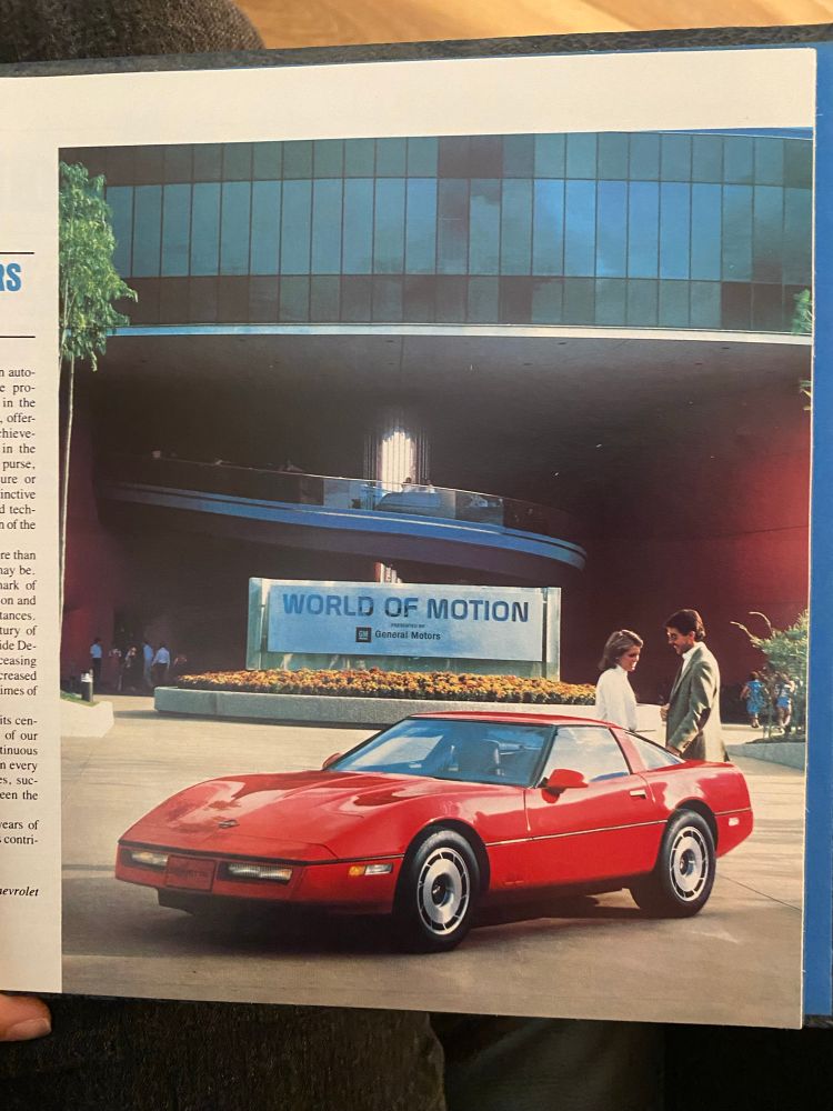 A photo from 1982-3 of a bright red GM car parked in front of the World of Motion with two very 80s looking people admiring it from behind it 