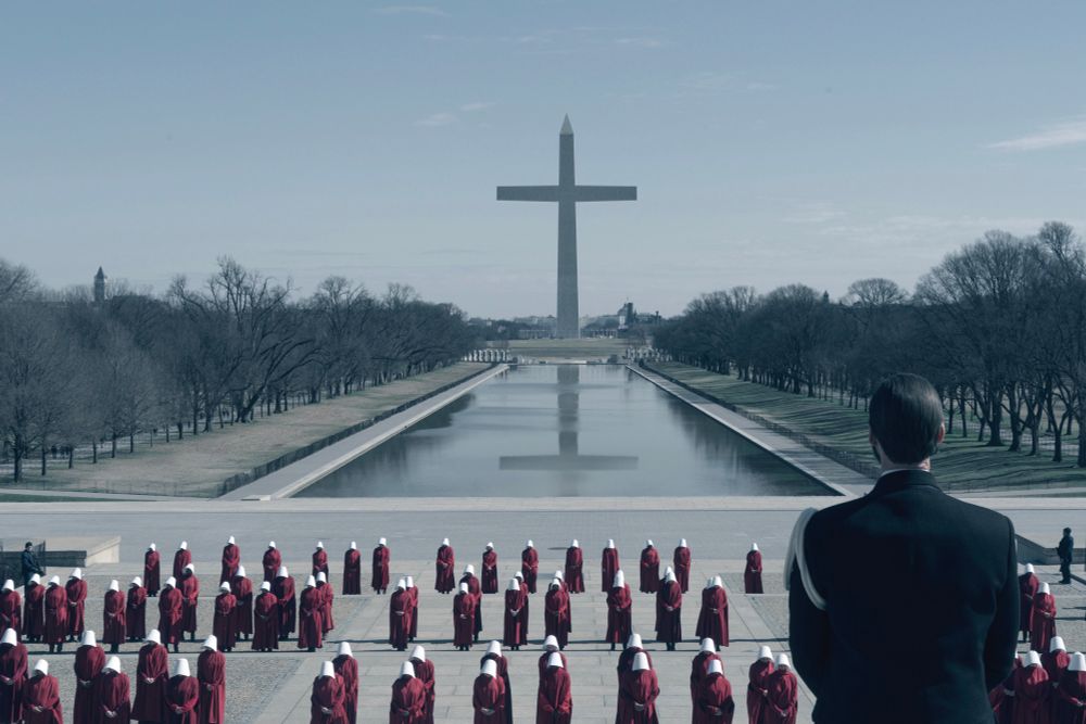 Scene from The Handmaid’s Tale. A Gilead general looks out over scores of handmaids lined up in military fashion, heads bowed. They are in front of reflecting pools in DC. In the background, the former Washington monument, converted into a giant cross looms large. 