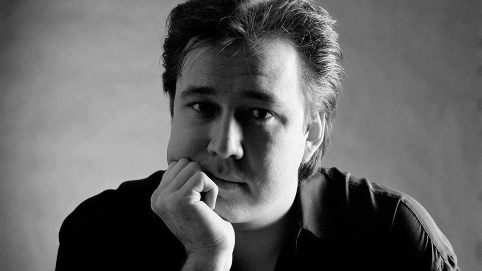 Bill Hicks, in black and white. He is posing towards the camera with his right hand curled up against the side of his face. 