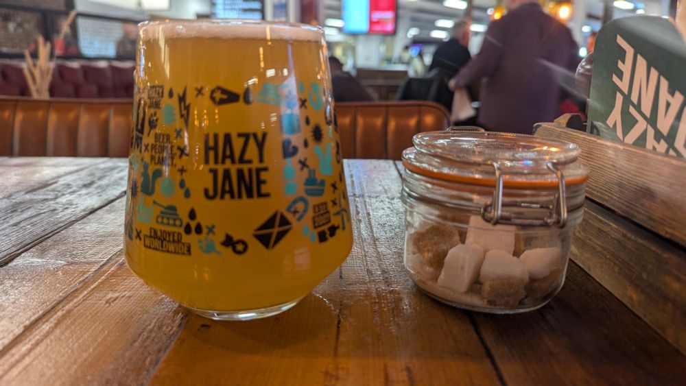 A pint of hazy Jane, with a well glass container of sugar next to it. Sitting on a wooden table. 