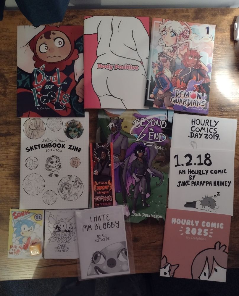 Photo of comics from Sequential Scotland 2025 from top left; A duel of fools by Cat Laird, Body Positive by Coin Operated Press, Demon Guardians by HesitantDoodle, Sketchbook Zine 2012 to 2015 by Ashling Draws, Beyond the End Volume 1 by Dusk Pendragon, Hourly Comics Day 2019 & Hourly Comic Day 2018 & Sonic the mini comic & Sonic 30th by Jake Parappa Hainey, I hate Mr Blobby by Ali Nytastic, and Hourly Comic Day 2025 by Delphine.