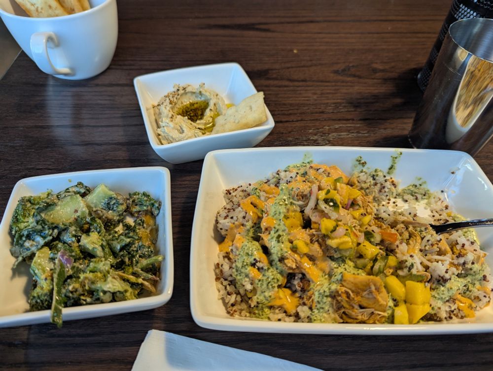 A plate of rice & quinoa, heaped with shredded chicken, mango salsa, and some green and orange sauces like the previously posted menu snip. A bowl of greens, a bowl with a dollop of hummus, and a mug of pita chips all sit next to the plate, 