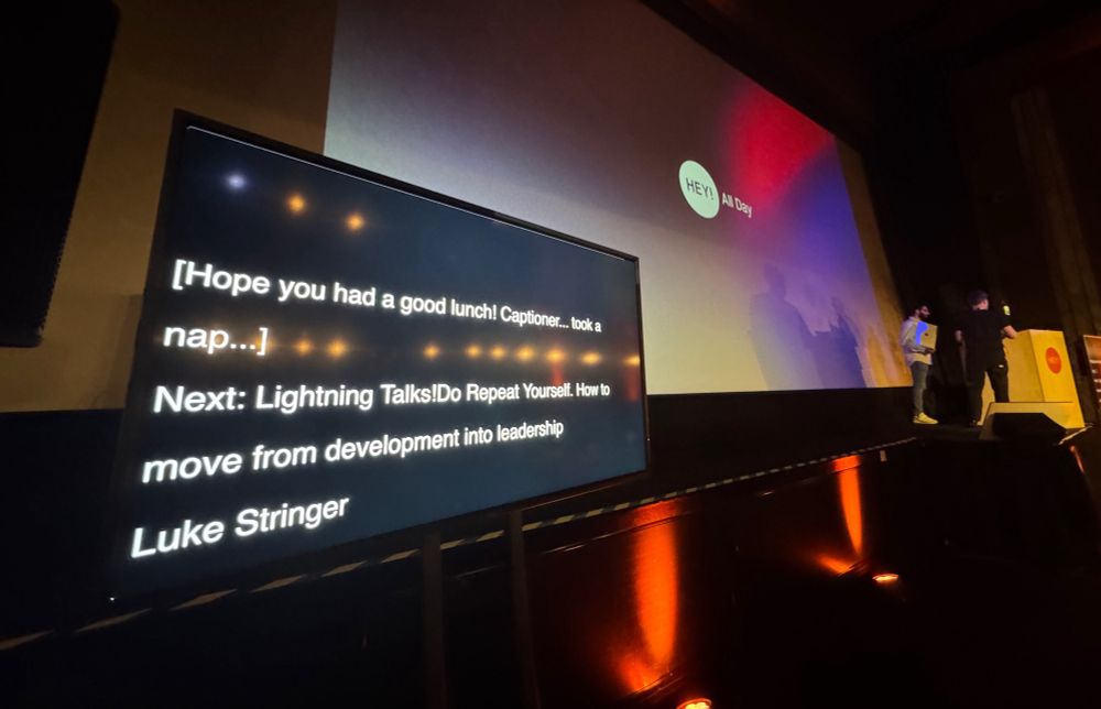 A screen at the front of a stage at a conference showing  live captioning. It reads, “[Hope you had a nice lunch… captioned took a nap]”
