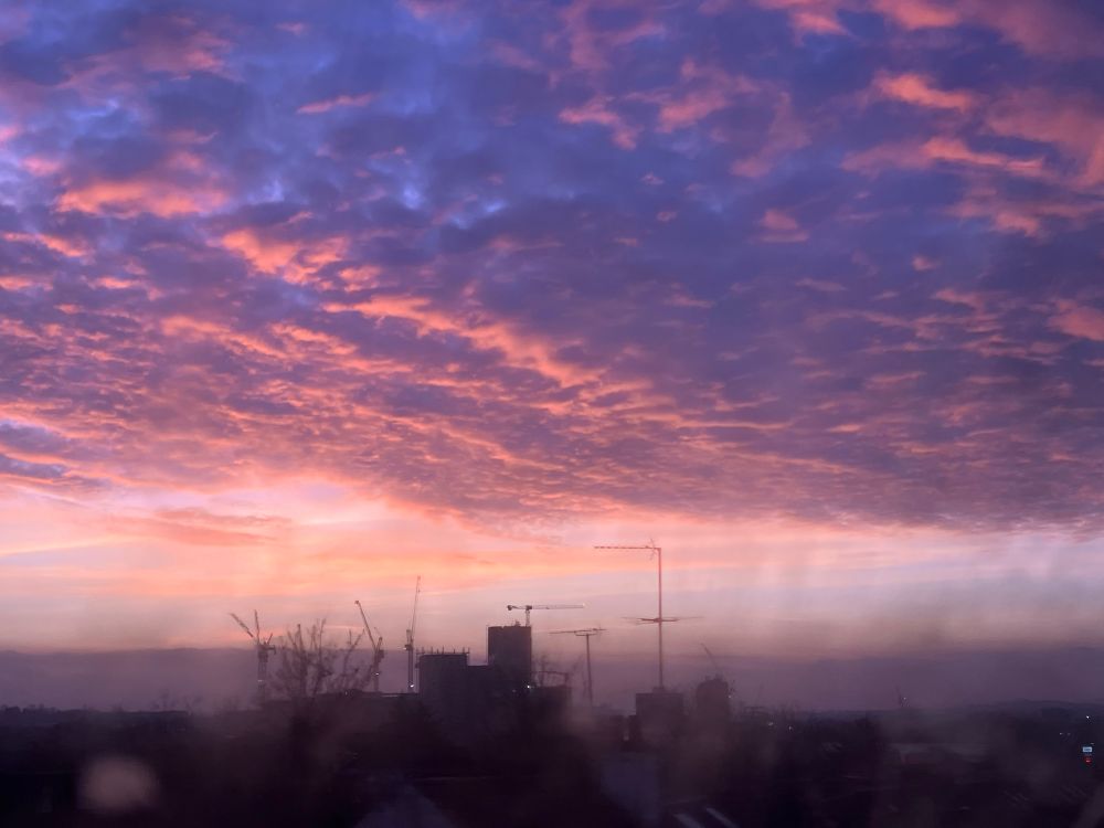Sunset through the clouds again - minutes later so less yellow and more pinks and purples - with buildings and cranes in mid ground. Taken from northern line tube train window 