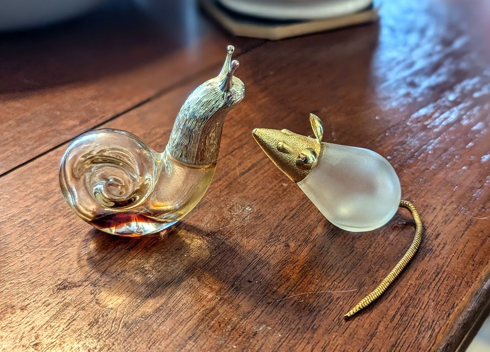 Photo of two small glass animals on a wooden table. The glass bottles are shaped like a transparent snail with a silver head. The second is an opaque glass mouse with a gold head and gold chain tail. Both bottles are empty. 