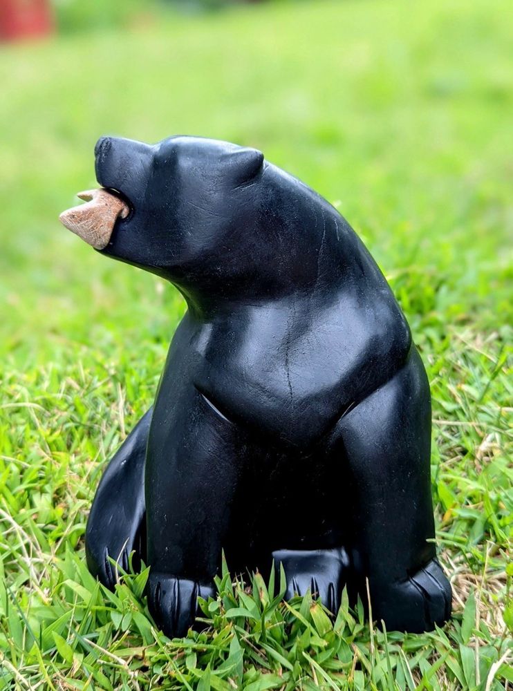 A face-on photo of a stone bear carving. The stone is solid black. The bear sits, head up and to the left. A small stone fish sits in its mouth, carved from a brown stone. The sculpture sits on a green grassy backdrop. 