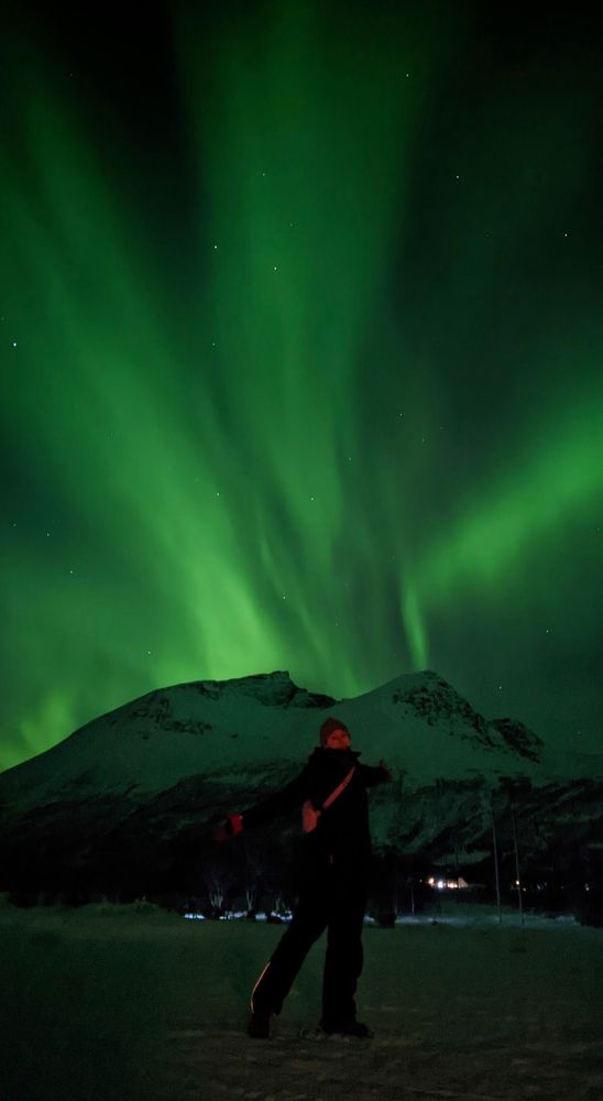 Photo of a silhouetted person standing in front of a snowy mountain, with vibrant green northern lights in the sky. 