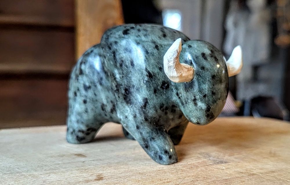 Photo of a small stone sculpture. The stone is blue with black spots, the shape resembling a buffalo walking forward, its horns made from roughly carved deer antler. 