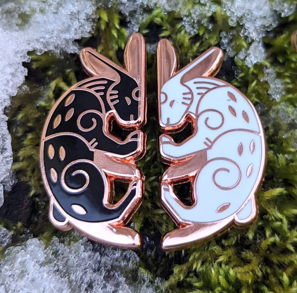 Photo of two enamel pins on a mossy background with some snow on the edges. The pins are a copper or rose pink metal, mirrored rabbit designs. Together they form an almost perfect circle, paws and foreheads meeting. The design is vector, almost Celtic looking. The left rabbit is black, the right white, their expressions serene. 