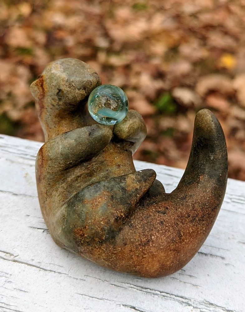A carving of a brown stone otter. It lays on its back, curled forward, tail up, two front libs holding a transparent green marble. 