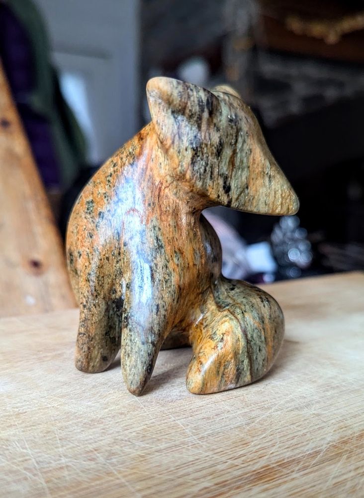 photo of a stone fox sitting on a wood base. The fox sits, glancing to the right, with two front legs showing and a fluffy tail. The stone is almost tiger stripe with grey-greens, blacks and oranges