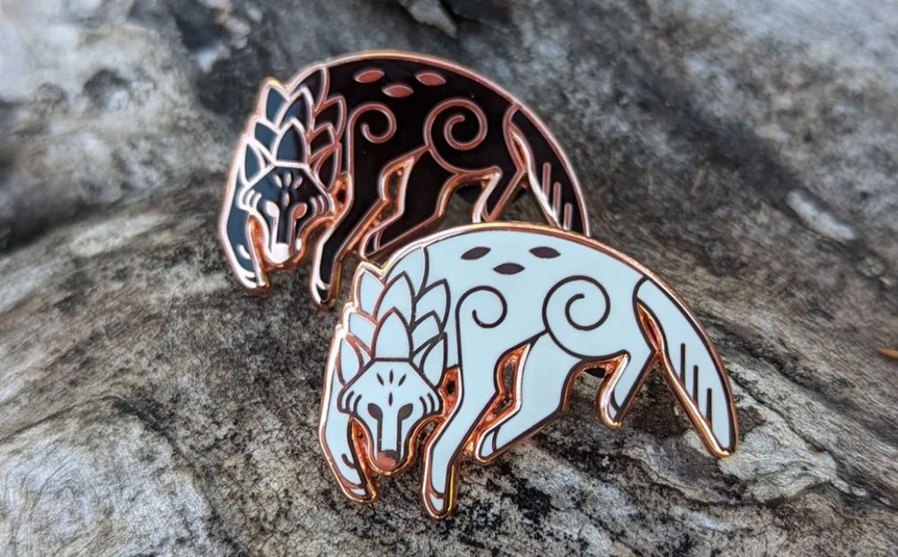 Photo of two enamel pins on a grey log. Pins are identical in design, with copper metal. The designs are of a wolf leaping to the left, head down, eyes on the viewer. One pin is in black, the other white. 