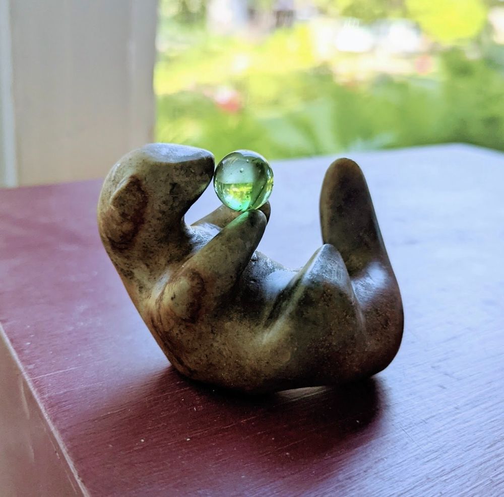 brown stone carving of an otter on its back, head, limbs and tail pointed up, body slightly curved. It holds a bright green marble in the front paws. It sits on a wooden red platform. 