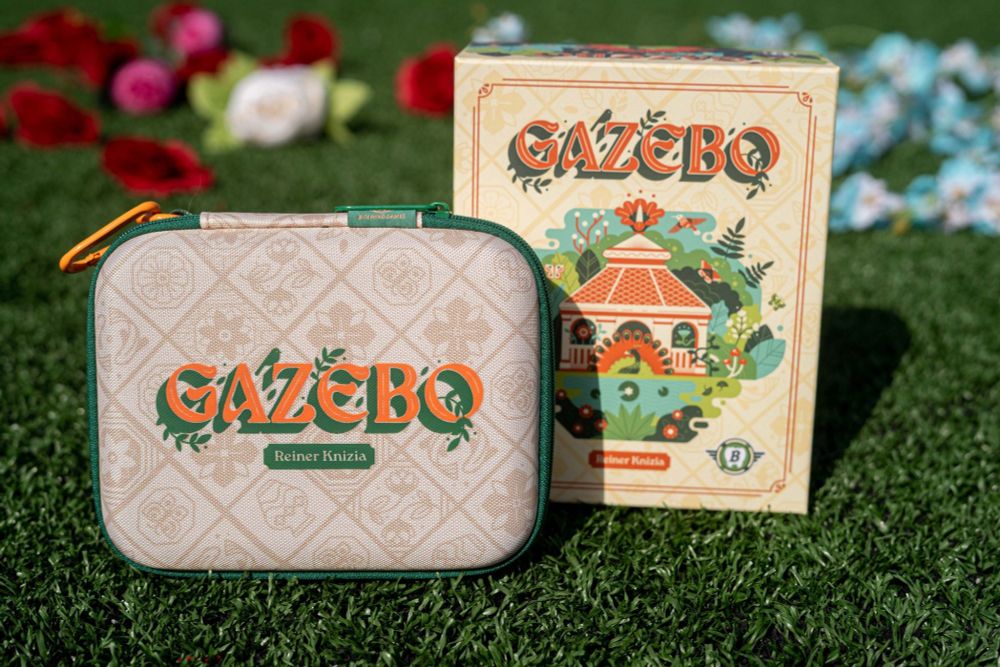 Photo of a board game box and fabric carrying case on a grassy background.  The case is a beige flower pattern with orange type that reads GAZEBO. The box is a brighter beige/yellow with a vector illustration of a gazebo, animals, and foliage in orange, green, dark greens and light blues. 