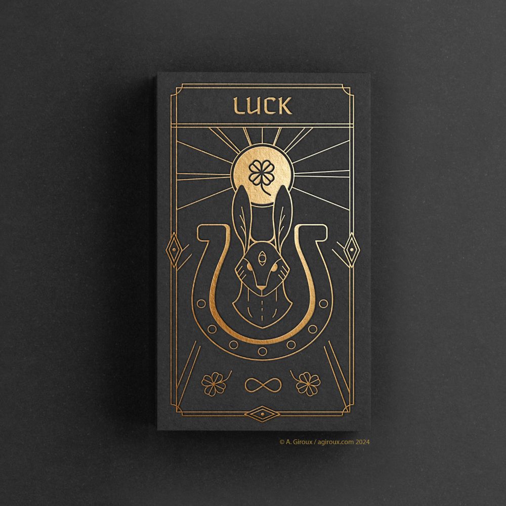 A tarot card on a flat black background. The card is a simple gold design printed on black paper. The top reads LUCK. A floating rabbits head stares at the viewer, ears straight, and a third eye is open on the forehead. It floats within a horseshoe. Above the head and between the hears is a sun disc with rays, and a four leaf clover sits within it. Below the illustrations are two more four leaf clovers with an infinity symbol between them. 
