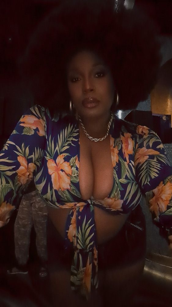 Fine ass Black woman in a floral tied crop top and jean shorts, a large Afro, and gold jewelry