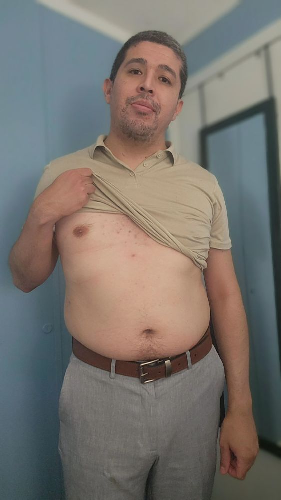 A man shows off his belly. He's wearing a beige polo and light grey slacks.
