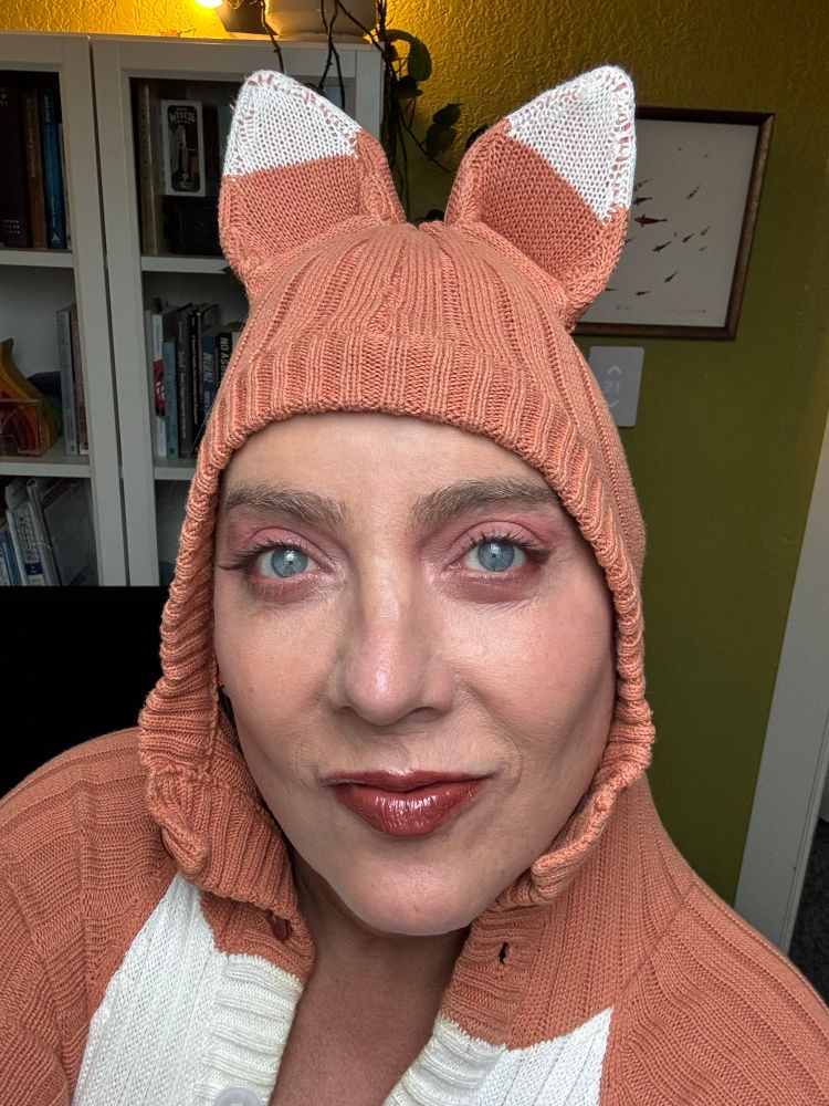 A white woman wearing a fox onesie/kigurumi smiles mischievously into the camera. 