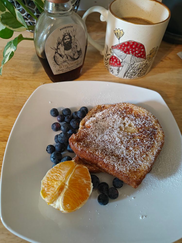 A white plate with French toast, blueberries, and an orange on it, with a mushroom themed mug and rum infused maple syrup in the background.