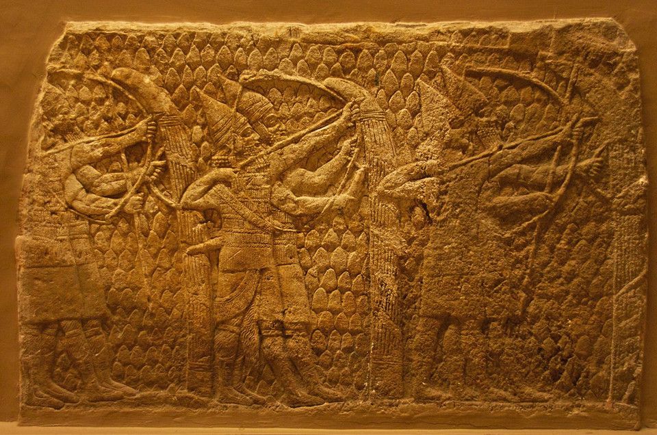 An image of a relief carved into stone showing three matched pairs of ancient Assyrian soldiers; each pair has an archer and a man holding a shield to protect him.