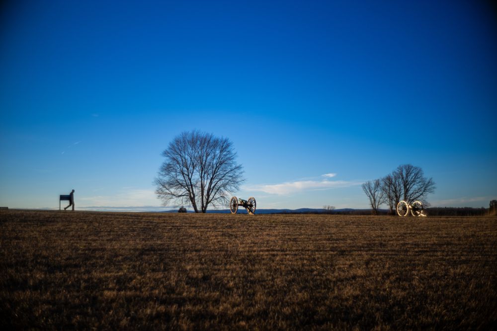 In the distance a silhouetted lone individual walks across a landscape towards civil war era artillery cannons in Manassas National Battlefield park.