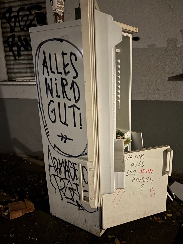 Ein alter, auf den Gehsteig gestellter Kühlschrank im Stadtteil Wedding, auf dessen Seitenwand behauptet wird, alles werde gut, während auf der Vorderseite die Frage gestellt wird, warum der Sohn betteln müsse.