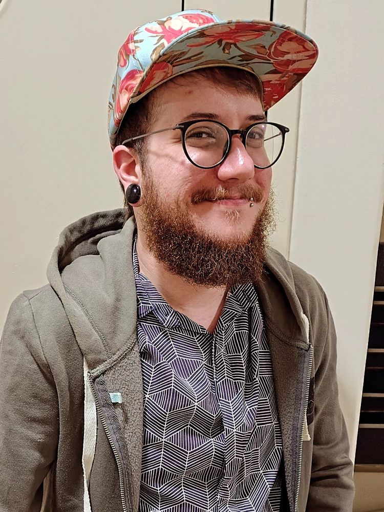 Foto minha com meio sorriso e uma barba consideravelmente cheia, olhando pra câmera. Usando óculos arredondados, um moletom de capuz cinza, uma camisa de botão com desenhos geométricos azul escuro e branco, um boné estampado de flores cor de rosa em um fundo azul claro.