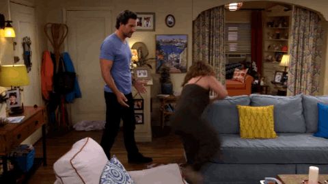 A GIF from Netflix's One Day at a Time featuring Max and Penelope. Penelope, in a matching brown tank top and cozy sweatpants, is jumping on a blue couch in her apartment so she can kiss the very tall Max, who is wearing a light blue t-shirt and jeans. 