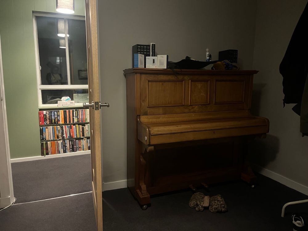 A piano in the hall with stuff on top of it, the door is open and books can be seen on a shelf 