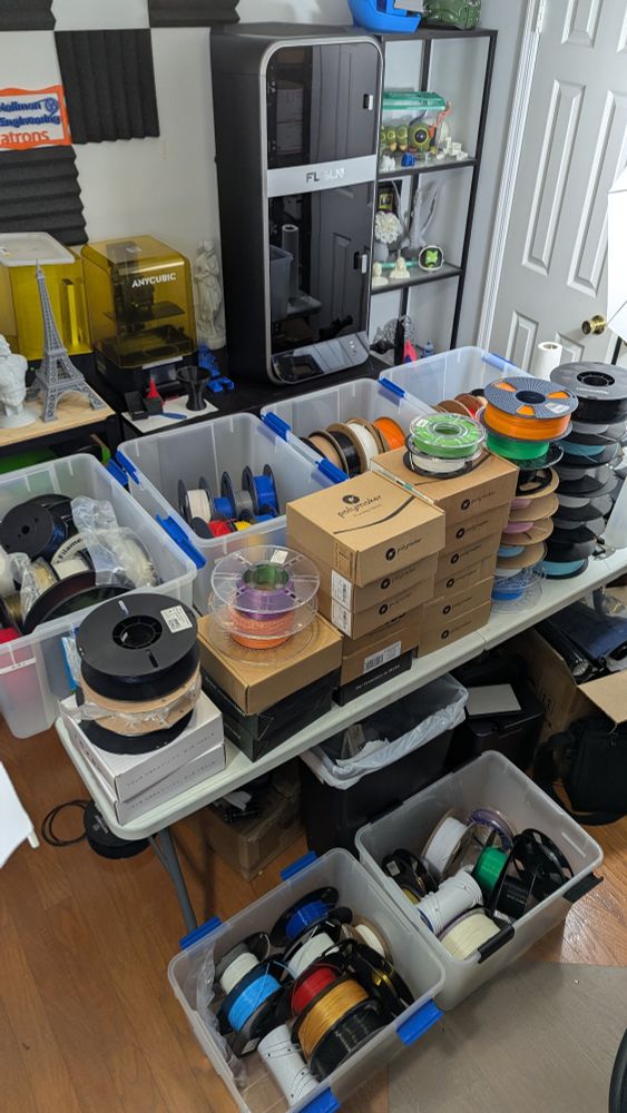 A workbench covered in an assortment of open and unopened spools of 3d printer filament, as well as four storage containers of filament. In front is two more containers of filament. 
