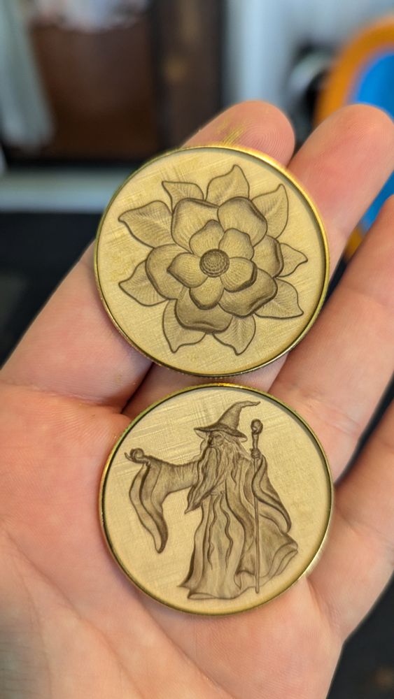 A hand holding two engraved brass coins. The top coin has a flower engraved into it. The bottom coin has a wizard holding a staff engraved into it. 