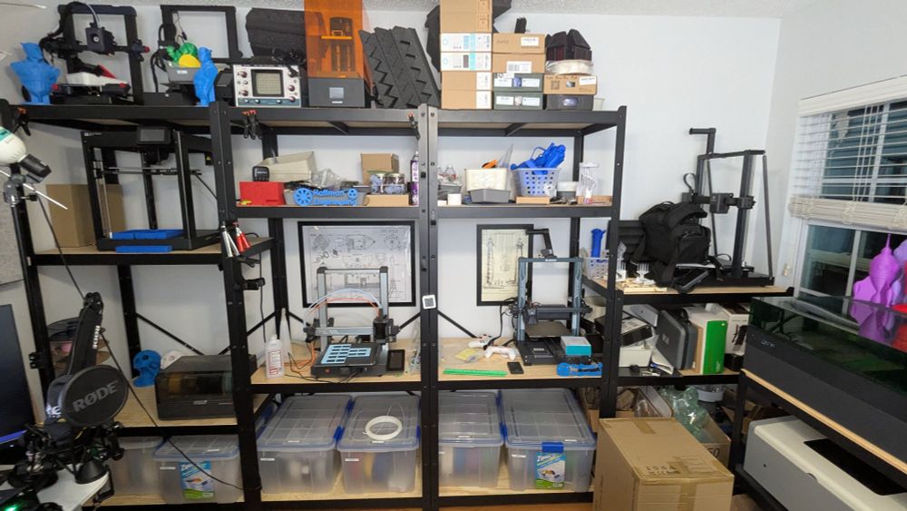 A wall of shelves. Bottom row contains six dry box storage containers of filament. The middle shelves contain two 3d printers. The top shelve contained a few more printers, as well as twelve Unopened boxes of filament. 