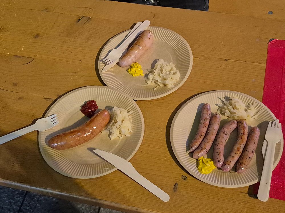 Bierbanktisch mit drei Papptellern, darauf zu sehen eine Frankfurter Wurst, eine Weißwurst sowie Nürnberger Rostbratwürstchen, jeweils mit etwas Sauerkraut sowie Ketchup oder Senf