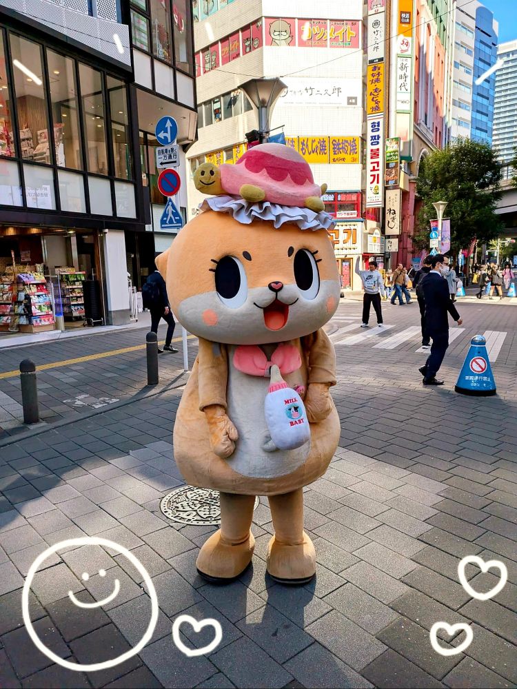 Chiitan (jap. Otterartiges Maskottchen) fotografiert auf dem Bürgersteig lässig stehend, im Hintergrund sind Hochhäuser und Geschäfte zu sehen.