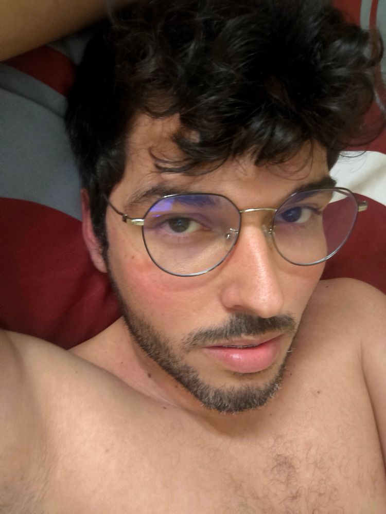 Selfie of me laying in bed, bare chest, wearing glasses, medium hair covering my forehead. I'm staring into the camera and my mouth is slightly less open than the previous picture.