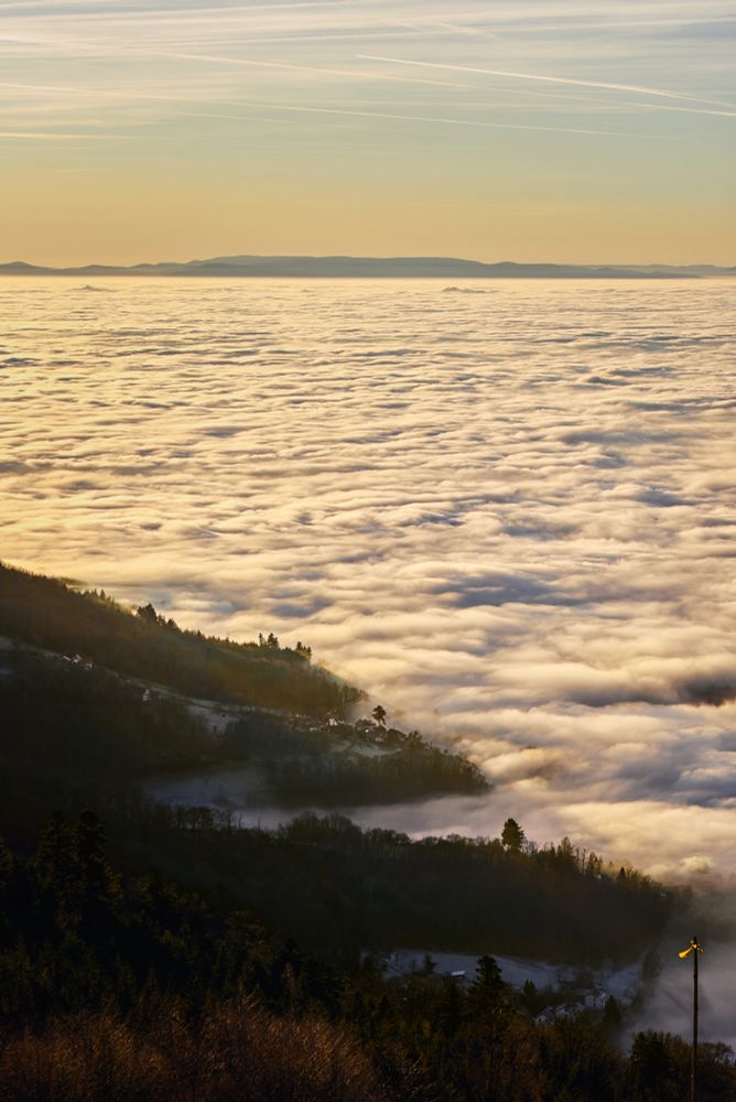 A landscape along a mountain range, gradually illuminated by the setting sun as it sinks into a sea of clouds.