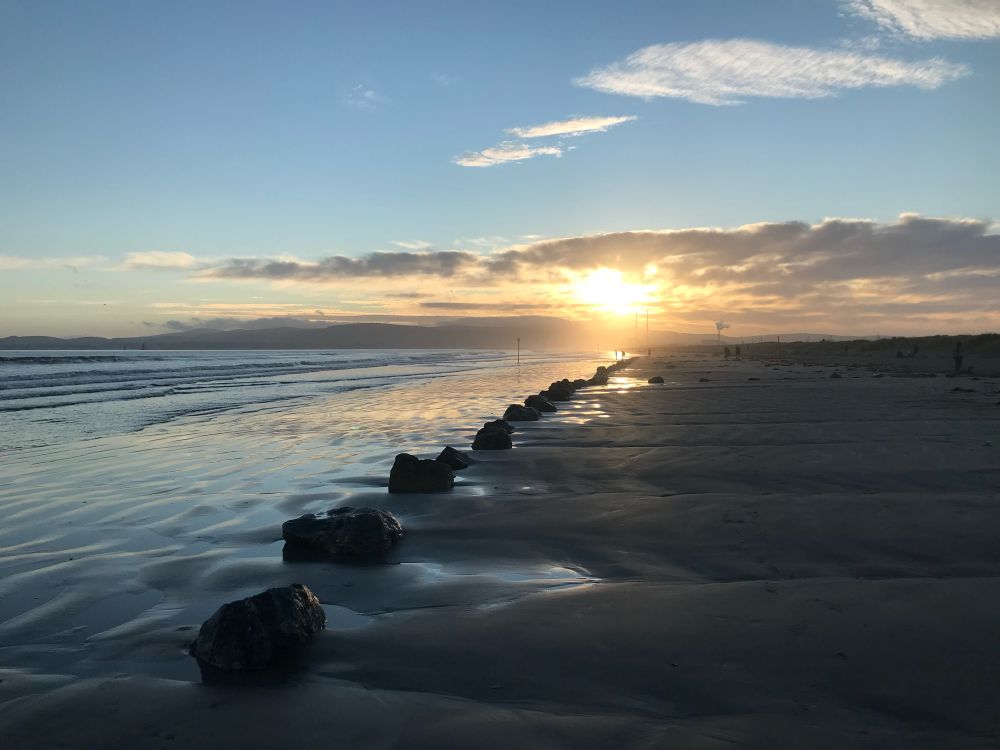 Dollymount strand, Dublin at sunset; sea to the left, sand to the right, sunsetting over Dublin mountains in the distance, blue sky with a little cloud